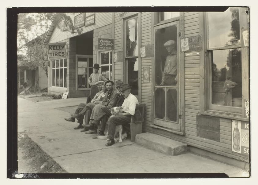 Men With Dog Sitting On Bench In Front Of General Store